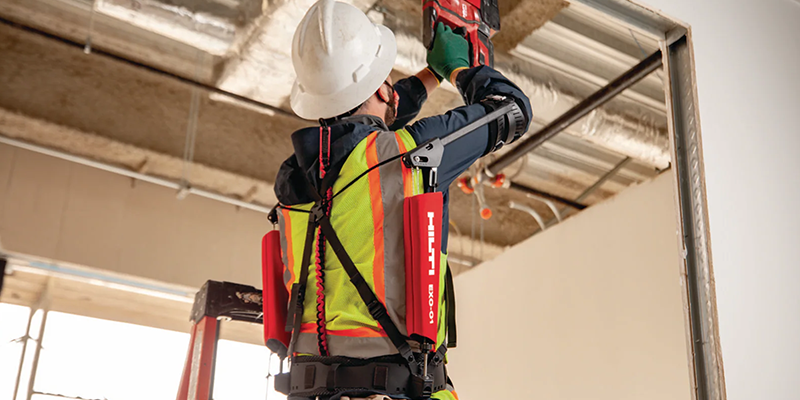 A person wearing the HA EXO 01 exoskeleton suit around their back as they work overhead at a construction site.